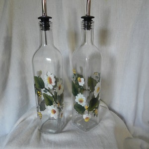 May include: Two clear glass bottles with silver spouts and black tops. The bottles are decorated with white daisies and green leaves.