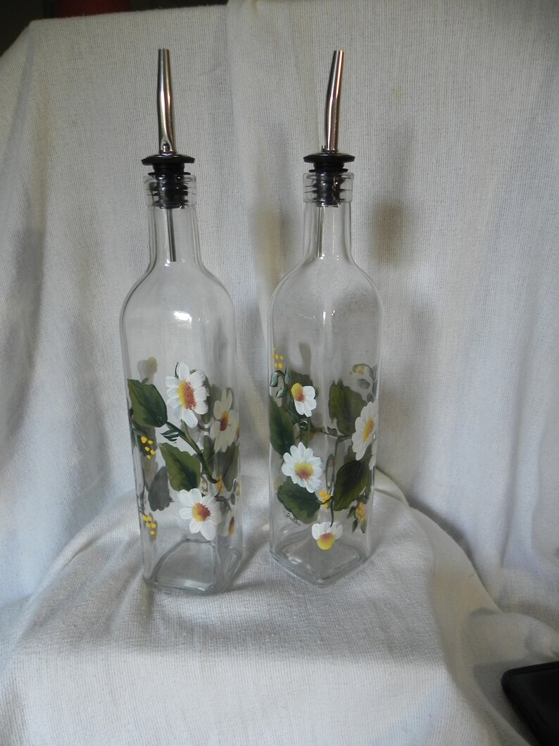 May include: Two clear glass bottles with silver spouts and black tops. The bottles are decorated with white daisies and green leaves.