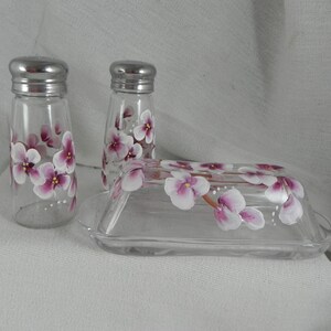 May include: A set of hand-painted glass salt and pepper shakers and a butter dish. The shakers and dish are decorated with pink and white flowers.
