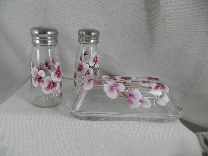 May include: A set of hand-painted glass salt and pepper shakers and a butter dish. The shakers and dish are decorated with pink and white flowers.