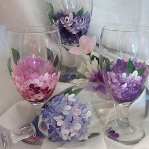 Puede incluir: Tres copas de vino pintadas a mano con diseños florales de color morado, rosa y azul. Las copas son transparentes y tienen un tallo y una base redonda.