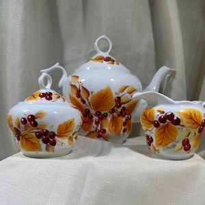 May include: A white ceramic tea set with a teapot, sugar bowl, and creamer. The set is decorated with a pattern of orange and yellow leaves and red berries. The teapot has a lid with a loop handle.