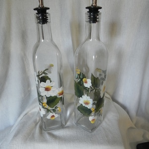 May include: Two clear glass bottles with black pour spouts and silver spouts. The bottles are decorated with hand-painted white daisies and green leaves.