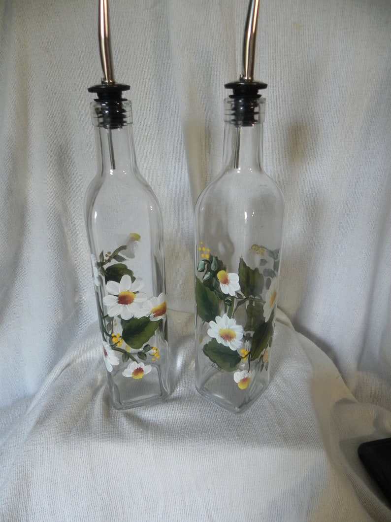 May include: Two clear glass bottles with black pour spouts and silver spouts. The bottles are decorated with hand-painted white daisies and green leaves.