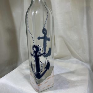 May include: A clear glass bottle with a black stopper and a painted design of two anchors and a rope. The bottle has a white and brown painted design at the bottom.