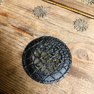 May include: A black, circular coaster with a textured surface and a decorative cut-out design. The coaster is placed on a wooden surface, with several small, metallic flower-shaped embellishments scattered around it. The coaster's design is visible.