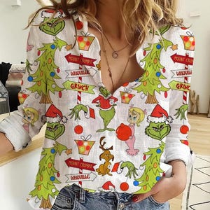May include: White button-down shirt featuring a festive Grinch-themed print. The design includes Grinch characters, Christmas trees, and directional signs with text. The long-sleeved shirt has a classic collar and buttoned cuffs, perfect for holiday wear.
