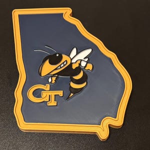 May include: A 3D-printed item in the shape of the state of Georgia, featuring a navy blue background and a yellow border. The image includes a yellow and black cartoon bee and the letters "GT" in yellow.