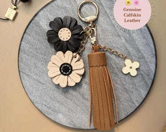 Könnte beinhalten: Ein Leder-Schlüsselanhänger mit zwei Blumenanhängern in Schwarz und Creme, einem braunen Quastenanhänger und einem goldfarbenen Kleeblatt-Anhänger. Der Schlüsselanhänger ist an einem goldfarbenen Ring befestigt. Der Text "Genuine Calfskin Leather" befindet sich in einem rosa Kreis.