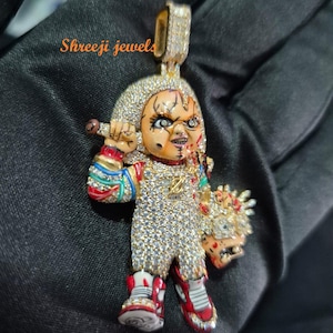 Puede incluir: Colgante dorado con forma de muñeco, incrustado con pequeñas piedras brillantes. El muñeco tiene la cara pintada, zapatos rojos y sostiene un cuchillo. Una pieza de joyería única.