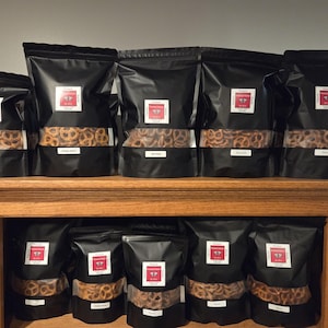 May include: Several black bags of pretzels are displayed on a wooden shelf. Each bag has a clear window showing the pretzels and a white label with red accents. The bags are arranged in rows.