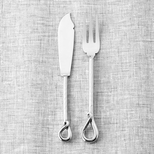May include: Silver butter knife and fork set. The knife has a curved blade and the fork has three tines. Both utensils have textured handles and teardrop-shaped end designs. The set is displayed on a gray textured surface.