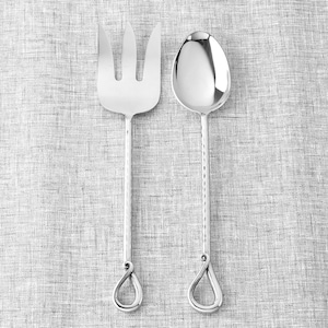 May include: A silver serving fork and spoon set. The fork has three tines, and the spoon has a rounded bowl. Both utensils have textured handles and teardrop-shaped loops at the ends. The set is displayed on a gray, textured surface.