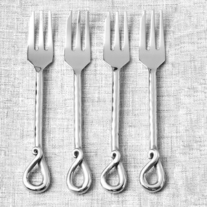May include: Four silver forks with a hammered texture on the handles and a decorative loop design at the base. The tines are evenly spaced, and the forks are arranged on a textured, light gray surface.
