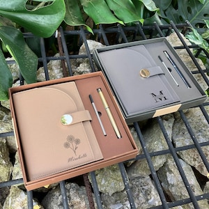 Op de afbeelding: Twee geschenkdozen, elk met een notitieboek en pen. Eén set heeft een tan notitieboek met een bloemenontwerp en de naam "Mirabelle". De andere heeft een grijs notitieboek met de initialen "M IVI". Beide sets bevatten een goudkleurige pen.