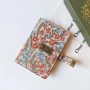 May include: A floral patterned journal with a combination lock. The cover features a beige background with red and blue flowers and green leaves. The journal has a strap with a metal clasp and a combination lock.