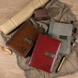 Op de afbeelding: Een assortiment lederen notitieboeken in bruin, rood en grijs, elk met een combinatieslot en decoratieve reliëfpatronen. Eén notitieboek is gegraveerd met de naam "Alice" en een vlinder.