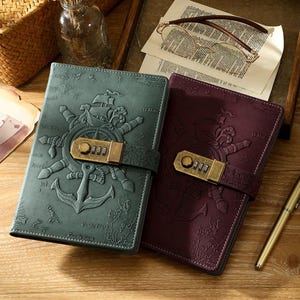 May include: Two leather journals, one teal and one burgundy, with embossed nautical designs. Each has a ship, anchor, and wheel motif, and a combination lock clasp. A pen and glasses are placed nearby on a wooden surface.