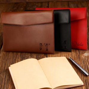 May include: Three leather document holders in brown, black, and red, with a notebook and pen on a wooden surface. The brown holder has the initials "JT" engraved, the black holder has the initial "T", and the red holder has the name "Odelia".