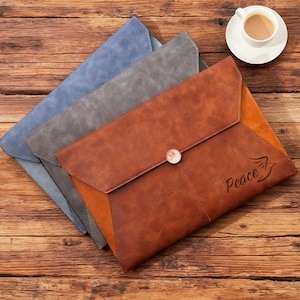 May include: Three leather document holders in blue, gray, and brown, arranged on a wooden surface. The brown holder has a button closure and the word "Peace" with a bird graphic. A cup of coffee sits in the upper right corner.