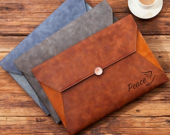 May include: Three leather document holders in blue, gray, and brown, arranged on a wooden surface. The brown holder has a button closure and the word "Peace" with a bird graphic. A cup of coffee sits in the upper right corner.