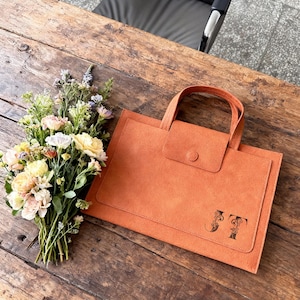 Può includere: Una borsa tote in camoscio arancione con una piccola patta e manico. La borsa presenta le iniziali "JT" in un carattere decorativo. Un bouquet di fiori di vari colori è posto accanto alla borsa su una superficie di legno.