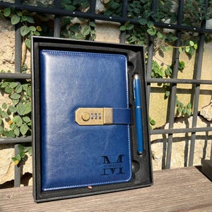 Op de afbeelding: Een blauw leren notitieboek met een combinatieslot en een bijpassende pen, gepresenteerd in een zwarte geschenkdoos. Het notitieboek heeft de naam "MARSHALL" gegraveerd aan de onderkant. De pen en het notitieboek hebben een bijpassende blauwe tint.