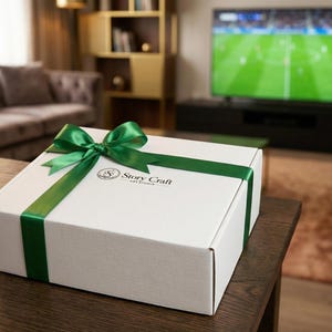 May include: A white gift box with a green satin ribbon and bow. The box has the text "Story Craft Art Studio" printed on the top. The box is sitting on a dark brown wooden table in a living room setting.