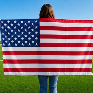 May include: A person holding an American flag with red and white stripes and a blue field of white stars. The flag is displayed against a backdrop of a green field and a clear blue sky.