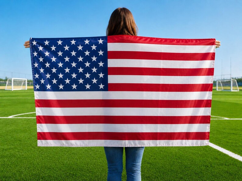 May include: A person holding an American flag with red and white stripes and a blue field of white stars. The flag is displayed against a backdrop of a green field and a clear blue sky.