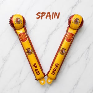 Puede incluir: Dos bastones de animaci&oacute;n inflables con los colores de la bandera espa&ntilde;ola, rojo y amarillo. Cada bast&oacute;n muestra el escudo de armas de Espa&ntilde;a y la palabra SPAIN en letras negritas. La palabra SPAIN tambi&eacute;n aparece en marr&oacute;n entre ambos.