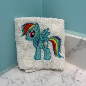 May include: A folded white terry cloth hand towel featuring an embroidered image of a cartoon pony with a rainbow mane and tail. The pony is light blue with a pink eye and wings. The towel is folded and resting on a white marble surface.