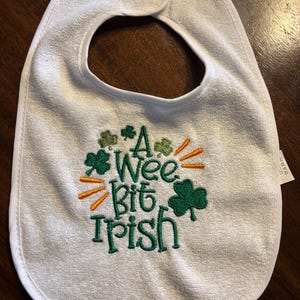 May include: White baby bib with green shamrocks and orange rays embroidered on it. The text "A Wee Bit Irish" is embroidered in green.