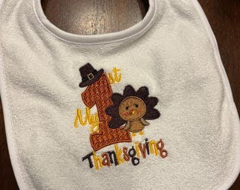 Puede incluir: Babero blanco para bebé con temática de Acción de Gracias. El babero presenta un diseño bordado con el texto "My 1st Thanksgiving" y un pavo de dibujos animados con un sombrero de peregrino. El babero tiene una abertura redonda para el cuello y un ribete blanco.
