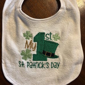 May include: White baby bib with green embroidery that says "My 1st St. Patrick's Day" with a shamrock and a leprechaun hat.