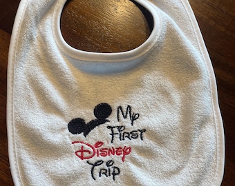 May include: White baby bib with a white trim. The bib has an embroidered Mickey Mouse head and the text "My First Disney Trip" in red and black thread.