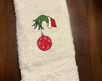 May include: White hand towel with an embroidered Christmas design. The design features a green hand holding a red ornament with white snowflake details. The towel is made of a soft, absorbent material.