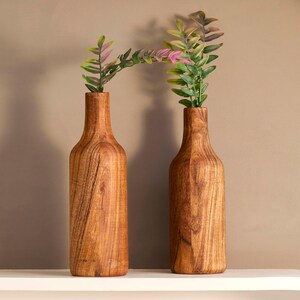 May include: Two wooden vases with a bottle shape, featuring a natural wood grain finish. Each vase holds a sprig of greenery with green and pink leaves. The vases are displayed on a white shelf against a neutral background.