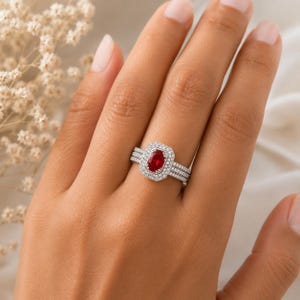 May include: A silver ring with a rectangular, deep red gemstone at the center, surrounded by a halo of smaller, clear stones. The ring has a triple-band design, with additional small stones set into the bands. The ring is on a hand.