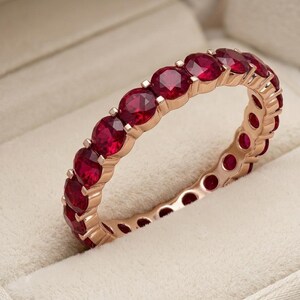 Puede incluir: Una sortija de tipo alianza en oro rosa, con una hilera continua de rubíes redondos de color rojo intenso engastados con garras compartidas. El anillo presenta un acabado metálico pulido y facetas de piedras preciosas muy brillantes.