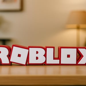 May include: A red and white Roblox logo displayed on a wooden surface. The letters are red with white block letters and a white square in the letter "O". The background includes a lamp and a plant.