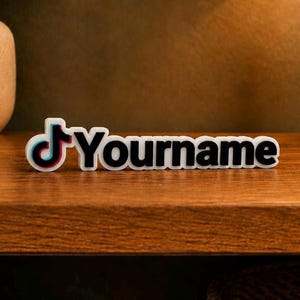 May include: A white and black desk sign with the TikTok logo and the custom text "Yourname". The logo features a musical note with red, blue, and black accents. The sign is displayed on a wooden surface.