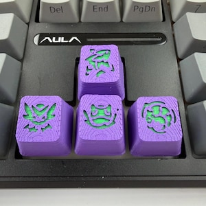 May include: Four custom purple keycaps with green accents, featuring carved designs of various characters. The keycaps are on a black keyboard, with the AULA logo visible. The keycaps are square and have a textured surface.