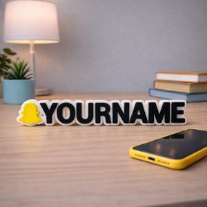May include: A white and black sign with the text "YOURNAME" in bold letters, accompanied by a yellow Snapchat ghost logo. A yellow smartphone is on the table. A lamp, plant, and books are in the background.