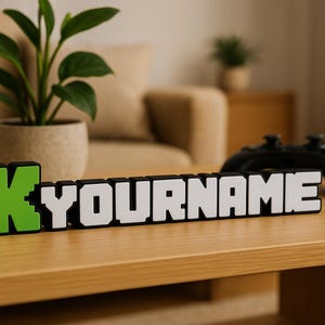 May include: A custom name sign with the text "K YOURNAME" in a pixelated font, with a green "K" and white letters. A black game controller sits on a wooden table, with a potted plant in the background.