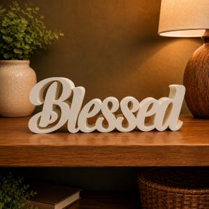 May include: Cream-colored, cursive "Blessed" wooden sign displayed on a wooden shelf. The sign is the focal point, with a warm, neutral color palette. The background includes a lamp, a vase with greenery, and a woven basket.