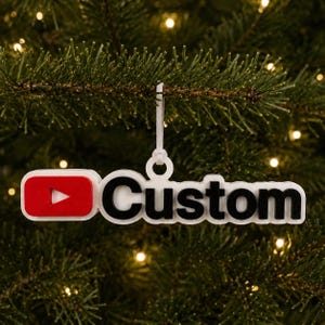 May include: A white hanging ornament featuring a red rectangular icon with a white play symbol on the left and the black text Custom on the right. The ornament has a circular loop at the top for hanging and is constructed with a layered design.