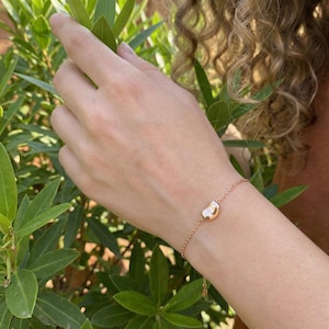 May include: A delicate gold-toned bracelet featuring a bean-shaped charm. The bracelet is shown on a wrist, with the charm positioned centrally. The chain is fine, and the bean charm has a smooth, polished surface. The background includes green foliage.