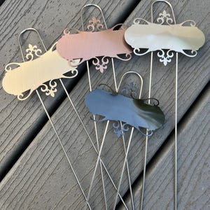 May include: A collection of five metal garden markers in various colors, including cream, rose gold, and silver. Each marker features a decorative flourish design and a long, thin stake for insertion into the soil. The markers are arranged on a wooden deck.