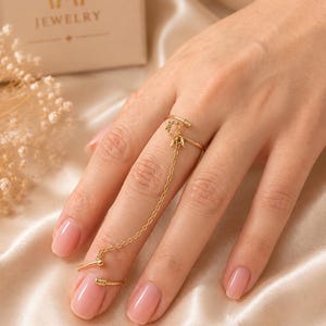 Puede incluir: Una pulsera de dedo dorada que presenta dos anillos abiertos conectados por una cadena delicada. El anillo superior muestra un pequeño motivo floral, mientras que el inferior es liso. Ambos anillos tienen extremos texturizados y enrollados.